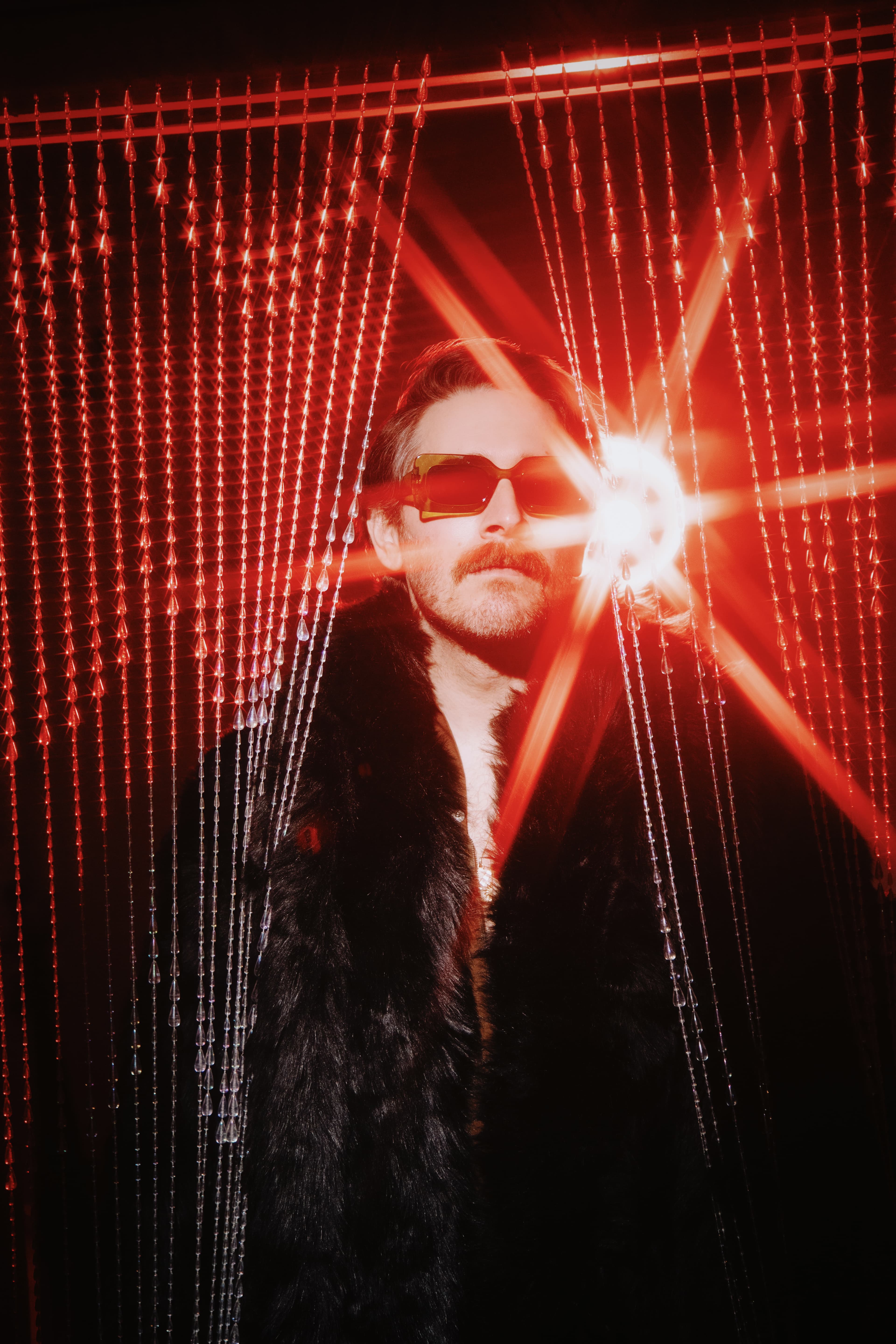 MGR Mike wearing sunglasses and a black coat beneath red lighting and a beaded curtain.